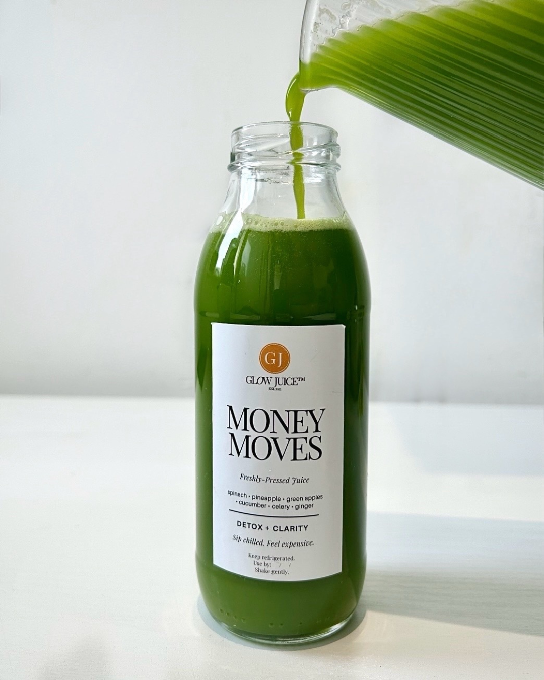 Green juice being poured into a bottle labeled 'Money Moves' with a white background.